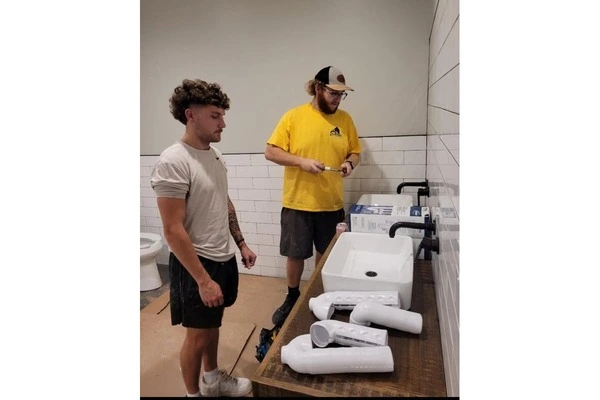 Two plumbers looking over white PVC drain fittings and sinks on a workbench